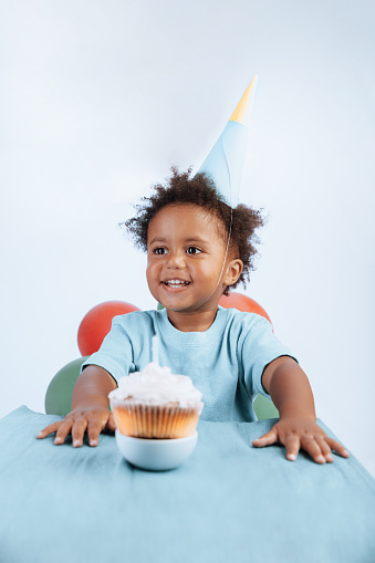 A small cheerfully smiling African-American boy dressed in a festive hat enjoys his birthday cake. A symbol of birthdays around the world.