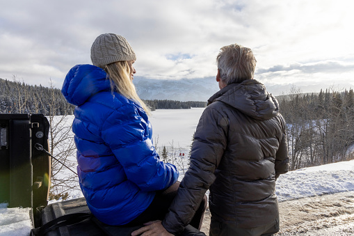 Mature couple lean against vehicle in winter and look across frozen lake and mountains covered in snow