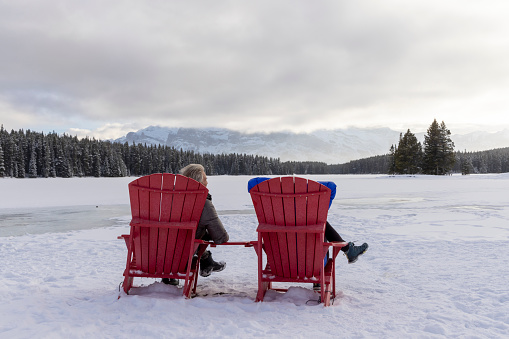 Mature couple lean against vehicle in winter and look towards foothills and mountains covered in snow