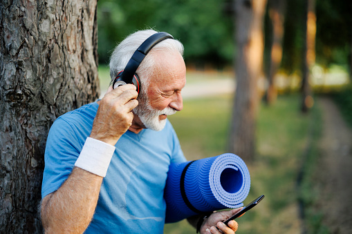 Portrait of sporty senior  man using phone, holding exercise mat and listening music outdoors