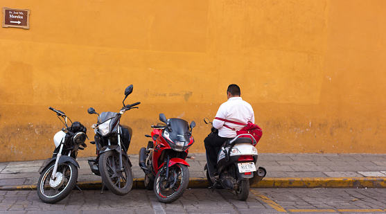 Oaxaca, Mexico: A man sitting on a parked motorbike against a vibrant orange wall in downtown Oaxaca.