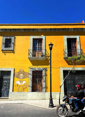 Oaxaca, Mexico: Two people ride a moped past a colorful building with traditional skeleton art in downtown Oaxaca.