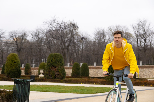 A cheerful young Caucasian man enjoys a leisurely bike ride through a park adorned with winter foliage, accompanied by people wearing yellow clothes, all reveling in the joy of cycling outdoors.