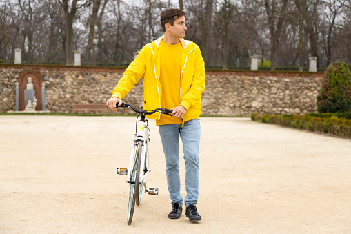 young Caucasian man, clad in a vibrant yellow raincoat, strolls with his bicycle, casting a sideways glance