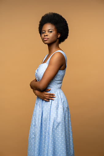 Peaceful African American woman looking at camera with confident glance, posing with arms crossed wearing in blue summer dress. Calm female isolated on studio beige background for advertisement
