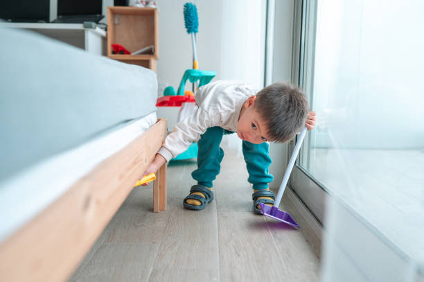 Two and a half year old boy joyfully sweeping the floor bellow the bed at home. Encourage early learning and responsibility with this heartwarming image of a little helper in action. A curious toddler sweeps away the floor with determination, showcasing his playful yet responsible nature while dressed in cozy clothing, bringing a sense of warmth and cleanliness to the home