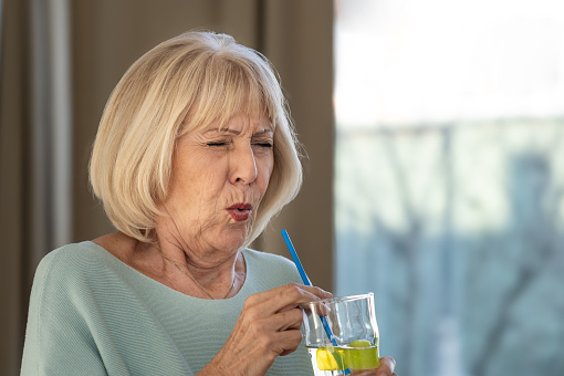A mature woman drinks a cold drink with ice through a straw and feels severe pain in her teeth due to damage to the tooth enamel. Hold onto the site of pain. She removes a straw and ice cubes from a glass of cold drink.