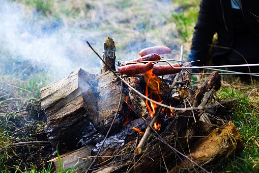 Traditional-style sausages roasting over a grassy bonfire with wood embers, emitting white smoke. Sausages have a beautiful golden-brown color.