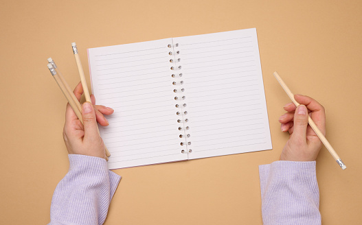 Open notebook with empty white sheets and a woman's hand holding a wooden pen on a yellow background, top view