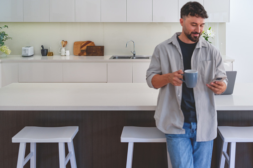 Man standing in the kitchen drinking coffee and looking at his mobile phone.