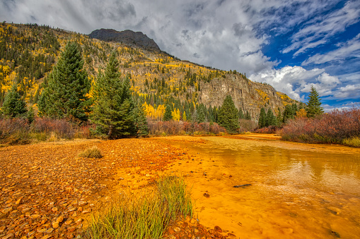 A bright orange stream caused by acid mine drainage with a mountain covered by yellow aspen trees on a sunny day near Ouray, Colorado