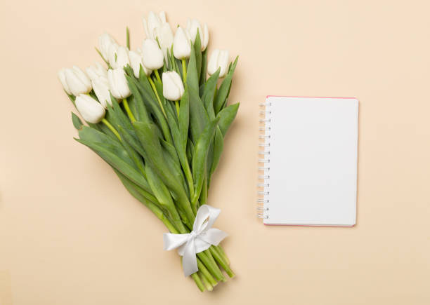White tulip bouquet with notebook on color background, top view