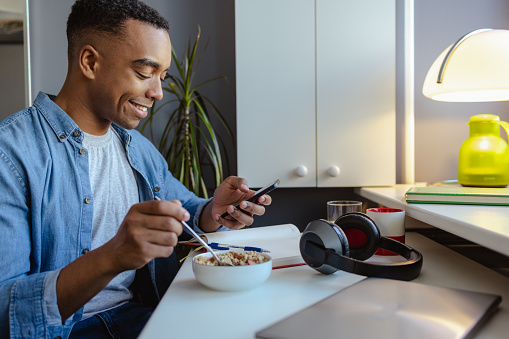 Young African American man home eating healthy breakfast and using mobile phone