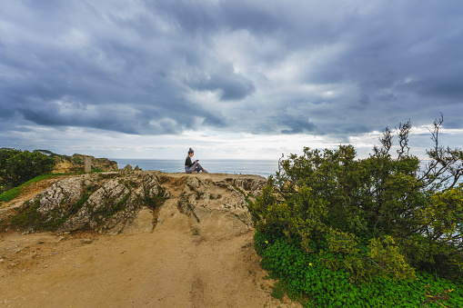 Algarve, Portugal, January 1, 2024. Young woman on a cliff overlooking a rugged coastline with arches and caves, surrounded by crashing ocean waves and a dramatic cloudy sky.