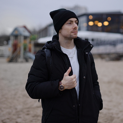 Young fashion man in black winter jacket and hat on the beach