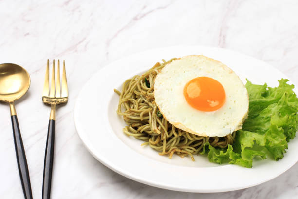 close up spicy healthy green instant fried noodle with shiny sunny side egg and lettuce on white ceramic round plate. - editorial front view sunlight light effect ストックフォトと画像