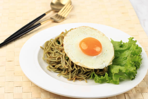 close up spicy healthy green instant fried noodle with shiny sunny side egg and lettuce on white ceramic round plate. - editorial front view sunlight light effect ストックフォトと画像