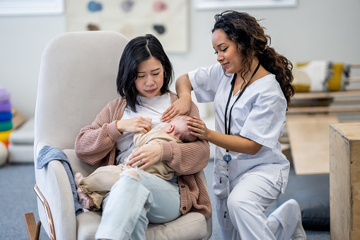 A new Mother attends a Lactation Consultant appointment as she looks for guidance with breastfeeding her daughter.  The practitioner is dressed professionally in scrubs as she calmly helps with positioning and latch.