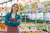 woman in apron keeping arms crossed and smiling in a greenhouse with many flowers