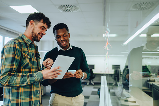 Portrait of a two diverse male colleagues writing their ideas for a new project on a colorful stickers glued on glass wall. Couple of multiracial businessman using paper sticky notes. Copy space.