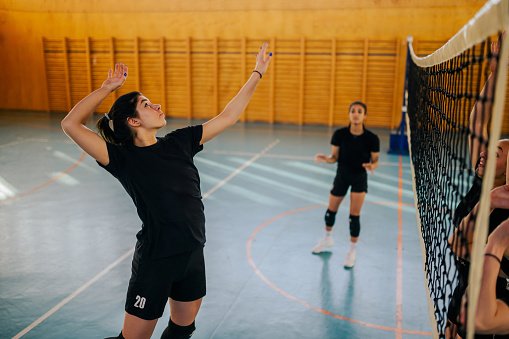 A teenage female volleyball team is practicing volleyball on court. A teenage sportswoman is hitting a ball over volleyball net on volleyball court and practicing with her interracial team. Copy space.