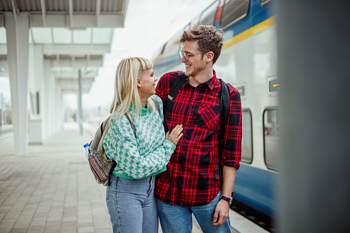 Portrait of two urban passengers standing near train at railroad station and hugging. Trendy passengers are about to enter the train at train station. Commuters hugging in front of a train at station.