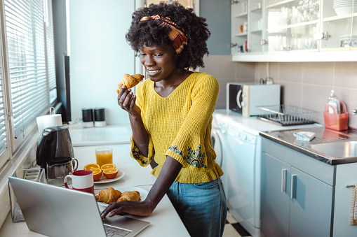 Photo of African American young woman using laptop and surfing the Net. She is having breakfast while working