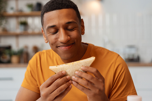 Portrait of positive African American man holding sandwich, eyes closed enjoying eating while sitting in kitchen at home. Concept of lunch, food, dinner