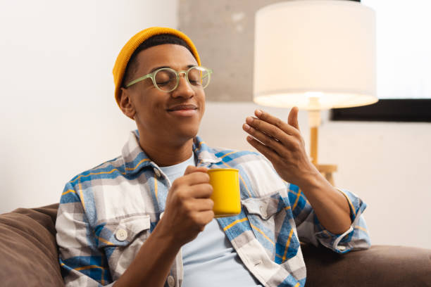 Happy African American man wearing yellow hat, stylish eyeglasses holding yellow cup, sniffing smell