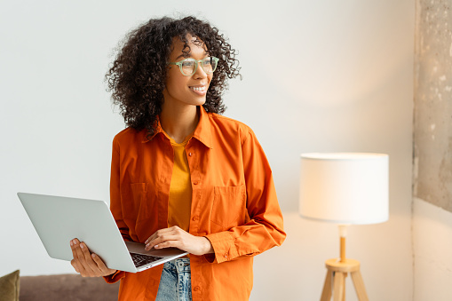 Smiling attractive African American woman, worker, manager holding laptop computer working online in modern office. Pensive freelancer wearing eyeglasses planning project looking away