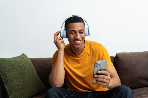 Portrait of smiling attractive African American man listening to music in headphones, holding mobile phone, using mobile app while sitting on sofa in room. Technology concept
