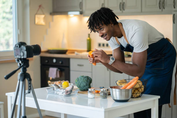 a multi-racial man bending down to look into a camera lens, recording his cookery vlog - camera lens home video camera broadcasting imagens e fotografias de stock