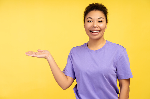 Portrait of smiling, beautiful African American woman wearing purple t shirt, holding hand on copy space, looking at camera standing isolated on yellow background. Advertisement concept