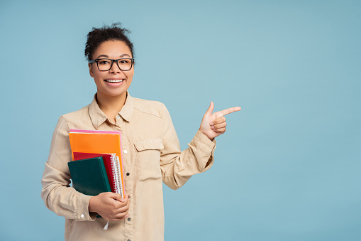 Happy beautiful African American woman businesswoman holding books wearing stylish eyeglasses pointing finger at copy space looking at camera isolated on blue background. Advertisement concept