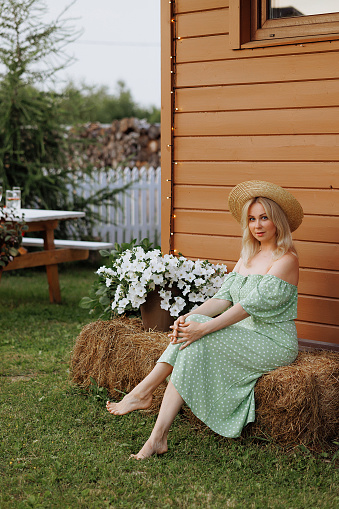 A young, beautiful woman in a green dress sits gracefully on hay bales in a rustic setting. The wooden house in the background. Concept: summer fashion photoshoot, rural relaxation.