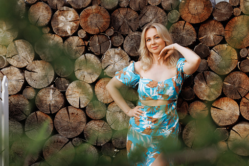 Confident adult woman in a vibrant, floral dress against a rustic wood log wall. Ideal for fashion and lifestyle content with copy space