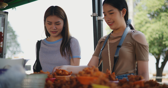 Two young Asian women selecting fish and chicken fried in plastic basket while buying meal in local food or somtum shop, tourist travel in countryside