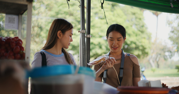 Two young Asian women selecting fish and chicken fried in plastic basket while buying meal in local food or somtum shop, tourist travel in countryside