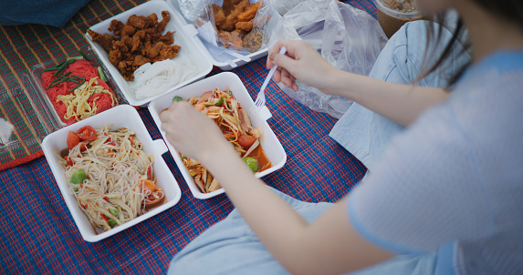 High angle view, Hands of Two young female tourists sitting on mat to eating the local food together, somtum, fried chicken and other food on lunch meal in park while tourist travel in countryside