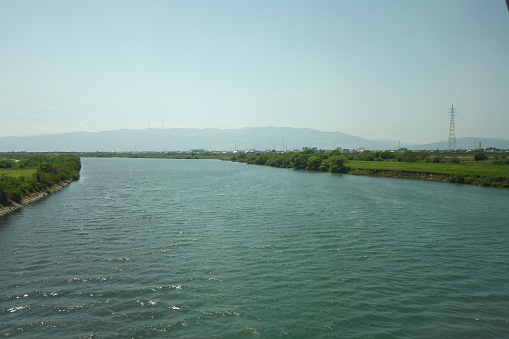 View of wide river in rural Japan with calm water surface and mountains and clear blue sky background. No people.