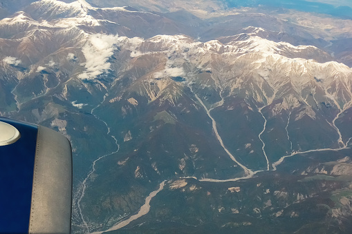 Shot of snowy mountains with mountain rivers descending over them from window of a passenger plane during the flight. Mountainous terrain, clouds, forests, all is an integral part of this landscape.