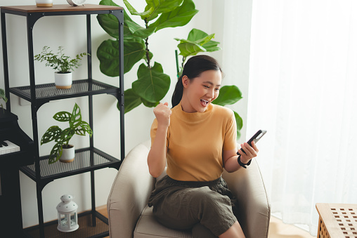 Asian woman holding and looking at smartphone happy excited and making a winning gesture sitting on armchair at home.