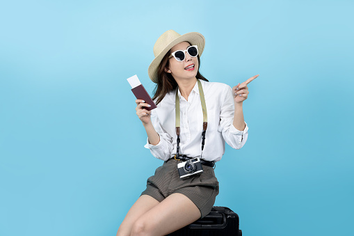 Happy smiling young Asian tourist woman sitting on luggage holding passport and hand pointing to copy space going to travel on holidays.