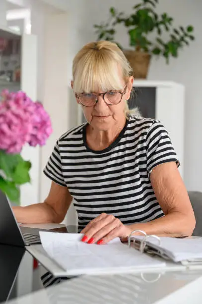 Focused mature woman in striped shirt and eyewear sitting at table while checking documents on paper during daytime at home