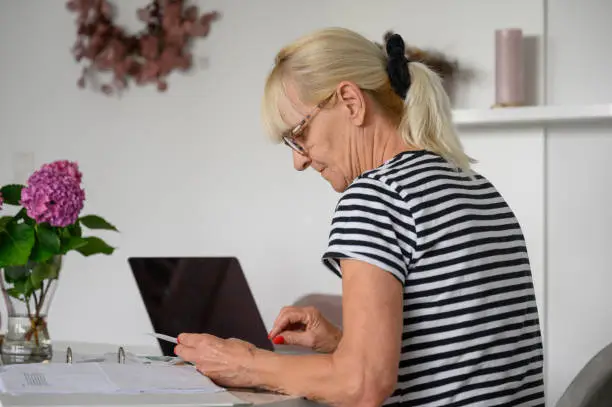Side view of senior blond woman, grandmother in casual shirt sitting at desk and checking bills while writing on notebook in modern living room at home daytime