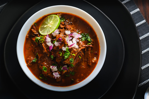 White bowl filled with delicious red Mexican beef birria stew on black plates. Served with red onion, cilantro and lime. Traditionally from Jalisco and central Mexican states.