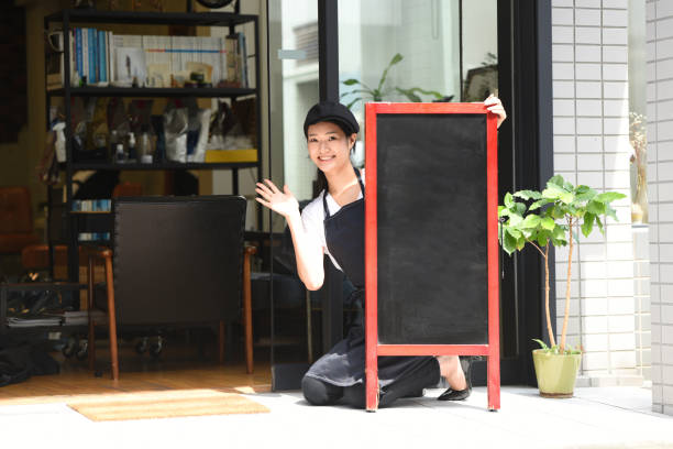 a young asian female clerk is setting up an easel sign in front of the restaurant to attract customers. - soda jerk 뉴스 사진 이미지