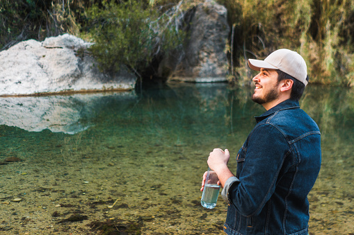 Smiling man with hat and a water bottle by a river.