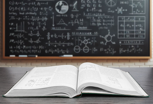 Open Textbook on Desk Against Blurred Blackboard With Mathematical Equations