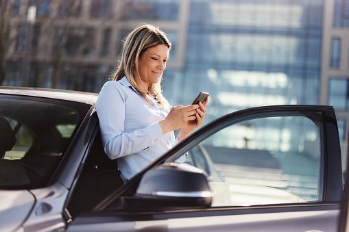 Young female entrepreneur reading a text message on smart phone while standing by her car on the street.